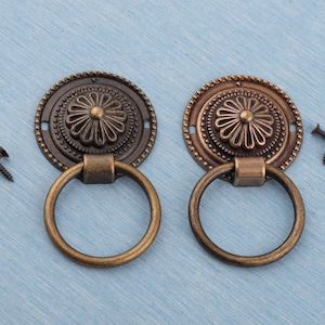 May include: Two antique bronze cabinet pulls with a floral design and ring handles. Each pull has a decorative circular backplate and includes mounting screws. The pulls are set against a light blue background.