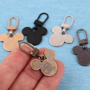 May include: Five metal zipper pulls shaped like mouse heads, each featuring a lobster claw clasp. The colors include rose gold, matte black, silver, dark bronze, and an antique brass finish with a small engraved logo on the front surface.