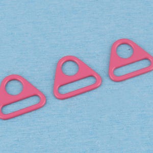 May include: Three pink metal triangle-shaped hardware components, each featuring a circular opening at the top and a horizontal rectangular slot at the base, designed for strap adjustment or attachment in various sewing and craft projects.