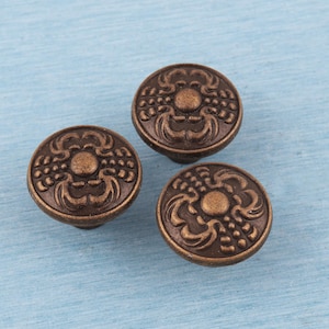 May include: Three bronze-colored cabinet knobs with a floral design. The knobs are round with a raised pattern of leaves and a central circular element. They are set against a light blue background.