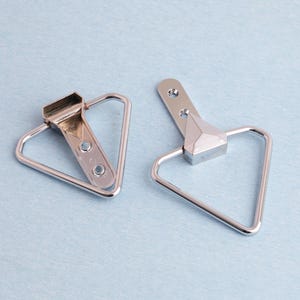 May include: Two silver picture frame hangers. Each has a triangular loop and a rectangular top with screw holes. The hangers are on a light blue surface.