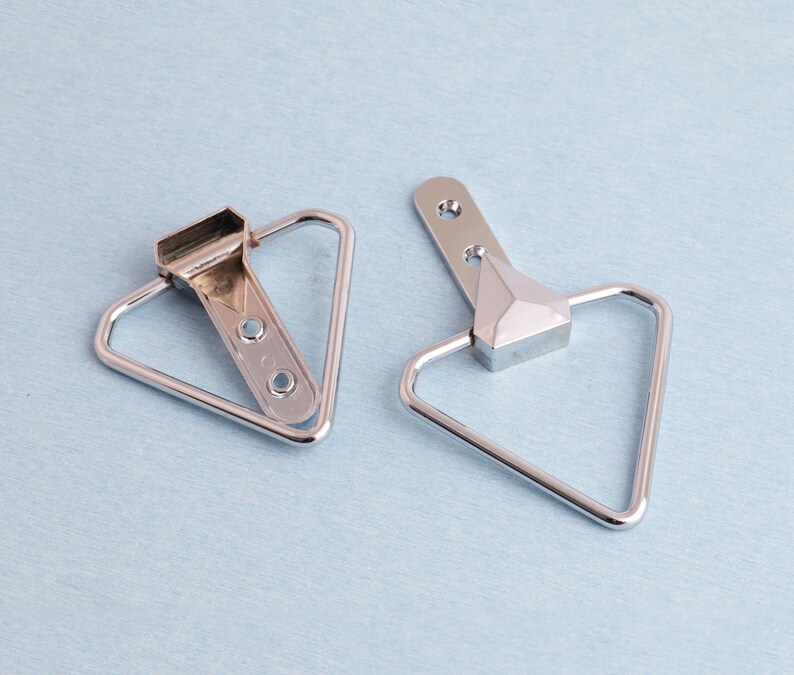 May include: Two silver picture frame hangers. Each has a triangular loop and a rectangular top with screw holes. The hangers are on a light blue surface.