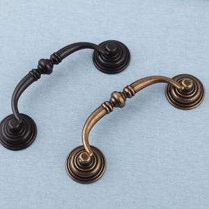 May include: Two decorative cabinet pulls. One is black, the other is bronze. Each pull has a curved handle and a circular base with concentric rings. The handles have a decorative, bulbous design.