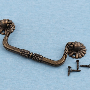 Könnte beinhalten: Antiker Bronze-Schubladengriff mit verzierten Details und einem gebogenen Griff. Der Griff wird mit vier Schrauben zur Montage geliefert.