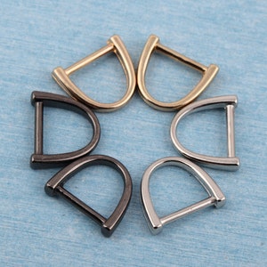 Puede incluir: Cinco anillas en D de metal en diferentes acabados: dorado, plateado y negro. Las anillas están dispuestas en círculo sobre un fondo azul.