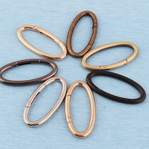 May include: An assortment of oval-shaped metal clasps in various colors, including gold, bronze, and black, arranged on a light blue surface. Each clasp has a hinged opening for attaching to other objects. The clasps are likely for use in jewelry or crafts.
