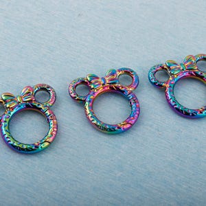 May include: Three iridescent metal charms in a mouse-ear shape with a bow on top. The charms have a circular ring and are in shades of blue, purple, green, and gold. The charms are on a light blue textured surface.