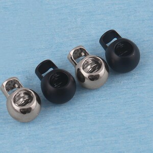 May include: Four spherical cord locks arranged in a diagonal line. Two of the cord locks have a polished silver-tone metallic finish, while the other two feature a matte black finish. Each cord lock includes a spring-loaded mechanism and a top loop.