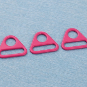 May include: Three bright pink, triangular-shaped plastic rings with a circular opening at the top and a straight bottom edge. The rings are arranged on a light blue, textured surface.