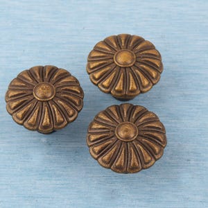 May include: Three antique bronze-colored cabinet knobs with a daisy-like design. Each knob features a central circular element surrounded by petal-shaped details. The knobs are set against a light blue, textured background, suggesting a vintage or rustic aesthetic.