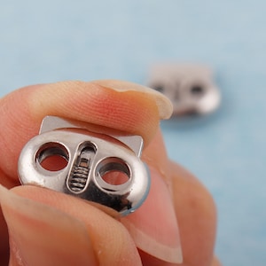 May include: A close-up of a silver-colored cord lock with a cat-ear design. The cord lock has two circular holes and a spring mechanism. Another cord lock is slightly out of focus in the background.