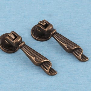 May include: Two antique bronze-toned metal drawer pulls featuring a circular mounting plate with a radial ribbed pattern and a hinged, tapered drop handle with vertical fluted detailing and a curved, flared end for furniture hardware.