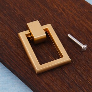 Op de afbeelding: Een goudkleurige rechthoekige trekker met een bijpassend rechthoekig bovenstuk. Naast de trekker zit een zilveren schroef. De trekker ligt op een donkerbruin houten oppervlak.