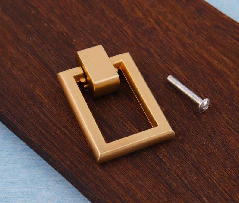 Op de afbeelding: Een goudkleurige rechthoekige trekker met een bijpassend rechthoekig bovenstuk. Naast de trekker zit een zilveren schroef. De trekker ligt op een donkerbruin houten oppervlak.