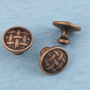 May include: Three bronze cabinet knobs with a woven basket design. Two are round with a knob base, and one has a flat, flower-shaped top. The knobs are set against a light blue background.