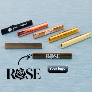 Könnte beinhalten: Eine Auswahl an Metall-Krawattenklammern in verschiedenen Farben: Schwarz, Kupfer, Gold und Bronze. Einige Clips zeigen das Wort "ROSE" mit einer Rose, andere "your name". Eine Logo-Option ist ebenfalls abgebildet.
