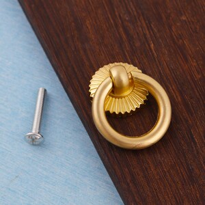 Op de afbeelding: Een goudkleurige kastgreep met een ringhandvat en een decoratieve, zonachtige achterplaat. Een zilveren schroef bevindt zich links van de greep. De greep bevindt zich op een donkerbruin houten oppervlak, met een lichtblauwe ondergrond.