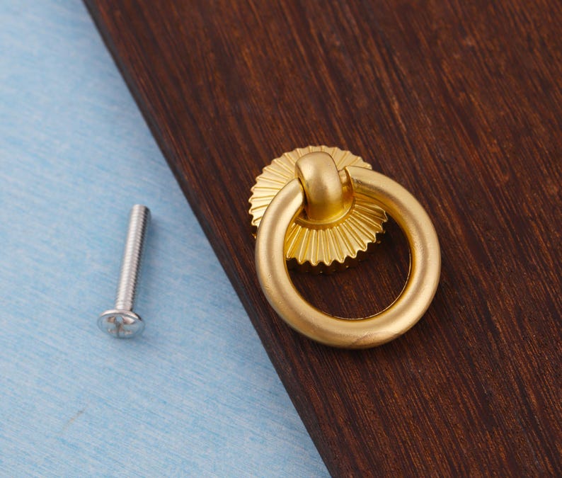 Op de afbeelding: Een goudkleurige kastgreep met een ringhandvat en een decoratieve, zonachtige achterplaat. Een zilveren schroef bevindt zich links van de greep. De greep bevindt zich op een donkerbruin houten oppervlak, met een lichtblauwe ondergrond.