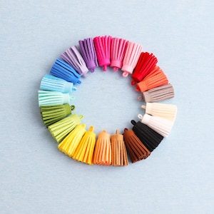 Puede incluir: Una disposición circular de borlas coloridas en varios tonos, incluyendo rosa, rojo, naranja, amarillo, verde y azul. Cada borla está hecha de un material suave y tiene una parte superior en forma de bucle. Las borlas están dispuestas sobre un fondo azul claro.