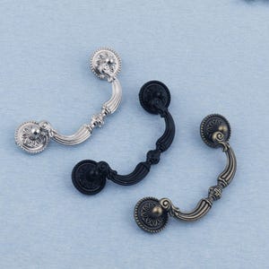 Puede incluir: Tres tiradores decorativos para cajones en plata, negro y bronce. Cada tirador presenta un diseño de roseta circular en los extremos y un asa curvada. La ferretería está sobre un fondo azul claro.