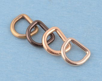 May include: Four D-rings arranged in a diagonal row, each featuring a distinct metallic finish. The set includes one matte brass, one dark gunmetal, one rose gold, and one polished light gold D-ring, all showcasing a smooth, rounded metal frame.