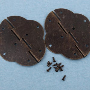 May include: Two antique bronze-coloured hinges with a four-petal design, each with a central hinge and multiple screw holes. Several small screws are scattered nearby, suggesting a set for furniture or craft projects.