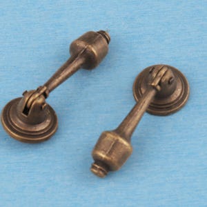 May include: Two antique brass-toned metal drop pulls for furniture. Each pull features a circular mounting plate with a hinged, elongated handle that terminates in a rounded, bulbous end. The finish has a darkened, weathered appearance.
