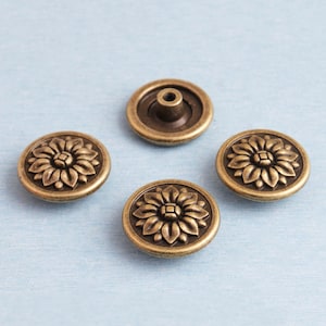 May include: Four antique brass-colored buttons with a floral design. Three buttons have a detailed flower pattern, while the fourth has a central post. The buttons are round and have a slightly raised design.