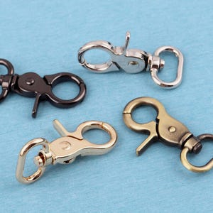 Peut inclure: Quatre mousquetons pivotants en métal avec différentes finitions, incluant argent brillant, noir mat, or poli et laiton antique. Chaque fermoir dispose d'un mécanisme à ressort et d'une boucle rectangulaire à la base pour la fixation.