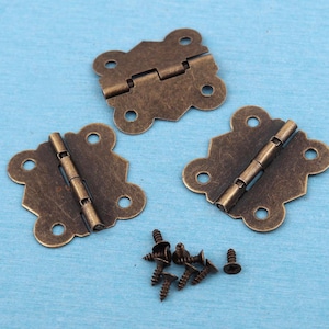 May include: Three antique bronze-colored metal hinges with a decorative scalloped edge design, each featuring four pre-drilled screw holes. A small pile of matching bronze-colored metal screws is positioned below the hinges for assembly.