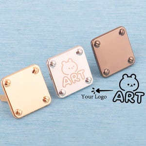 May include: Three square metal cabinet knobs in gold, silver, and rose gold. Each knob has four screws in the corners. The silver knob has a bear graphic and the word "ART". The background is a light blue wood grain.