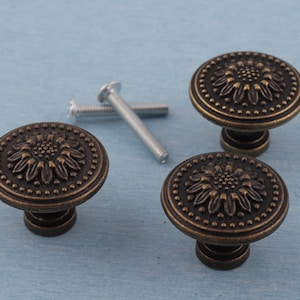 May include: Three antique bronze cabinet knobs with a sunflower design and a dotted border. Two silver screws are also visible. The knobs have a dark bronze finish and are likely made of metal.