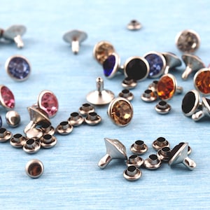 Puede incluir: Una colección de remaches metálicos plateados con centros de piedras preciosas facetadas de colores. Las piedras preciosas son de varios colores, incluyendo rojo, rosa, azul, ámbar y negro. Pequeños ojales metálicos a juego también están dispersos.
