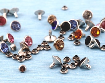 May include: A collection of silver-toned metal rivets with colorful, faceted gemstone centers. The gemstones are in various colors, including red, pink, blue, amber, and black. Small, matching metal grommets are also scattered around the rivets on a light blue surface.