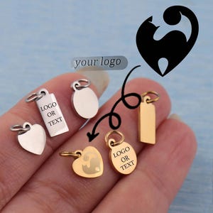 Puede incluir: Una colección de pequeños colgantes metálicos de varias formas, incluyendo corazones, óvalos y rectángulos. Algunos colgantes son de color plateado, mientras que otros son dorados. Los colgantes tienen pequeños bucles para sujetarlos a la joyería. Algunos colgantes tienen el texto "LOGO OR TEXT".