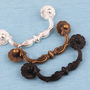 Könnte beinhalten: Drei dekorative Schubladengriffe in Silber, Bronze und Schwarz. Jeder Griff hat ein Blumendesign an den Enden und einen gebogenen Griff. Die Griffe sind aus Metall und werden auf einer hellblauen Oberfläche präsentiert.