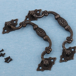 May include: Two antique bronze cabinet handles with ornate floral designs and decorative screw heads. The handles have a curved shape and are approximately 5 inches long, perfect for adding a vintage touch to furniture.