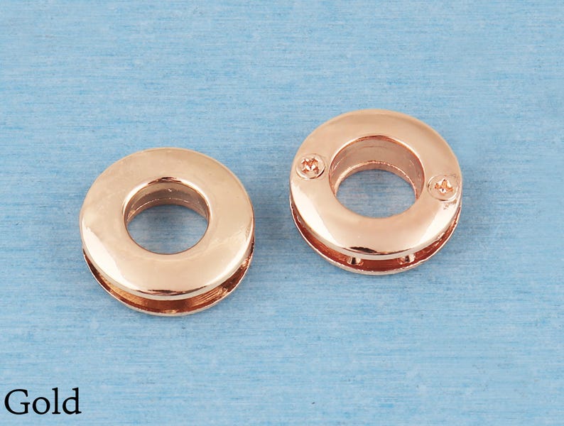 May include: Two circular gold-toned metal grommets with a smooth, polished finish. Each grommet features a central opening and a flat, ring-shaped design. The right grommet displays two small, visible screw heads on its surface. The word Gold is printed.