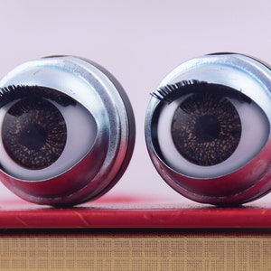 Puede incluir: Dos ojos de muñeca grandes, redondos, plateados con pestañas negras e iris marrones. Los ojos están sobre una superficie roja.