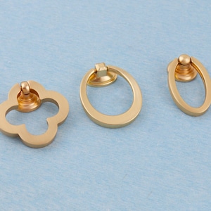 May include: Three gold-coloured cabinet pulls in different shapes: a four-leaf clover, an oval, and a smaller oval. Each pull has a ring handle and a circular base. The pulls are displayed on a light blue surface.