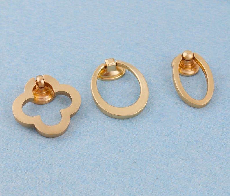 Może przedstawiać: Trzy złote uchwyty do szafek o r&oacute;żnych kształtach: czterolistna koniczyna, owal i mniejszy owal. Każdy uchwyt ma uchwyt pierścieniowy i okrągłą podstawę. Uchwyty są wyświetlane na jasnoniebieskiej powierzchni.