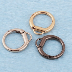 May include: Three round metal clasps in gold, rose gold, and dark bronze. Each clasp has a hinged opening and a small loop. The clasps are arranged on a light blue surface.