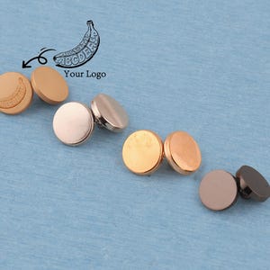 May include: Four pairs of circular metal buttons in varying metallic finishes including matte gold, rose gold, polished gold, and dark gunmetal. One button features a small engraved banana graphic with the letters ABCDE. Includes a banana logo graphic.