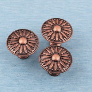 May include: Three bronze-colored cabinet knobs with a floral design. Each knob features a central circular element surrounded by petal-shaped details. The knobs are set against a light blue wooden surface.