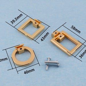 Op de afbeelding: Drie goudkleurige kastgrepen in verschillende vormen: een vierkant (43 mm x 39,5 mm), een rechthoek (58 mm x 36 mm) en een cirkel (42 mm x 40 mm). Inclusief montageschroeven. De hardware wordt weergegeven op een lichtblauwe ondergrond.