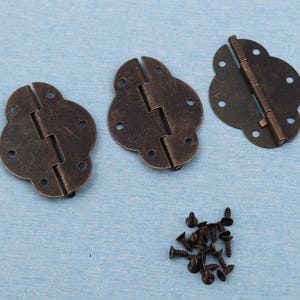 Könnte beinhalten: Drei antikfarbene Scharniere in Bronze mit dekorativer Kleeblattform und ein Haufen kleiner, passender Schrauben. Die Scharniere sind geöffnet und geschlossen, wodurch der Mechanismus sichtbar wird. Ideal für Vintage-Projekte.