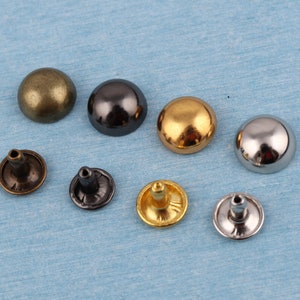 May include: Assortment of round metal rivets in various finishes, including gold, silver, bronze, and black. The rivets have a domed top and a flat base with a post for attaching to fabric or leather.