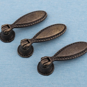 Puede incluir: Tres tiradores de cajón de bronce antiguo con diseño ovalado y bordes decorativos. Cada tirador presenta una base redonda y un asa con un diseño detallado y ornamentado. La ferretería se encuentra sobre un fondo azul claro.