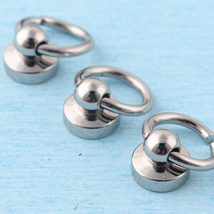 K&ouml;nnte beinhalten: Drei silberne Metall-Schnapphaken mit runder Basis. Die Haken sind an einem runden Ring befestigt.