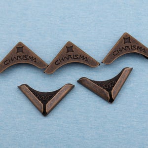 Könnte beinhalten: Fünf antike bronzefarbene Metallecken. Jede Ecke hat eine gebogene Kante und das Wort "CHARISMA" ist oben eingraviert. Ein sternförmiges Design befindet sich über dem Wort. Ideal für Scrapbooking und Bastelarbeiten.